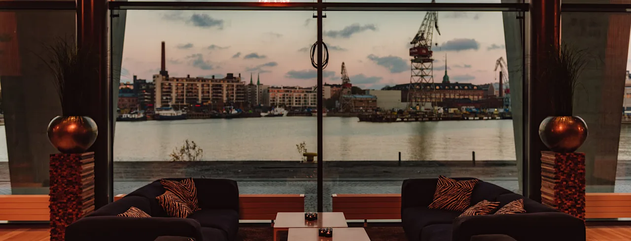 En moderne lounge med mørke sofaer og mønstrede puder vender mod et stort vindue, der viser en havneudsigt med bygninger, kraner og en solnedgangssky, mens fyrværkeri lyser op horisonten.