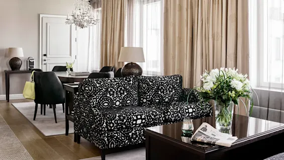 Elegant, mønstret sofa står i en elegant stue med en lysekrone ovenover, omgivet af lyse gardiner og mørke møbler. En avisoverskrift lyder "LIFE & ARTS" på bordet.