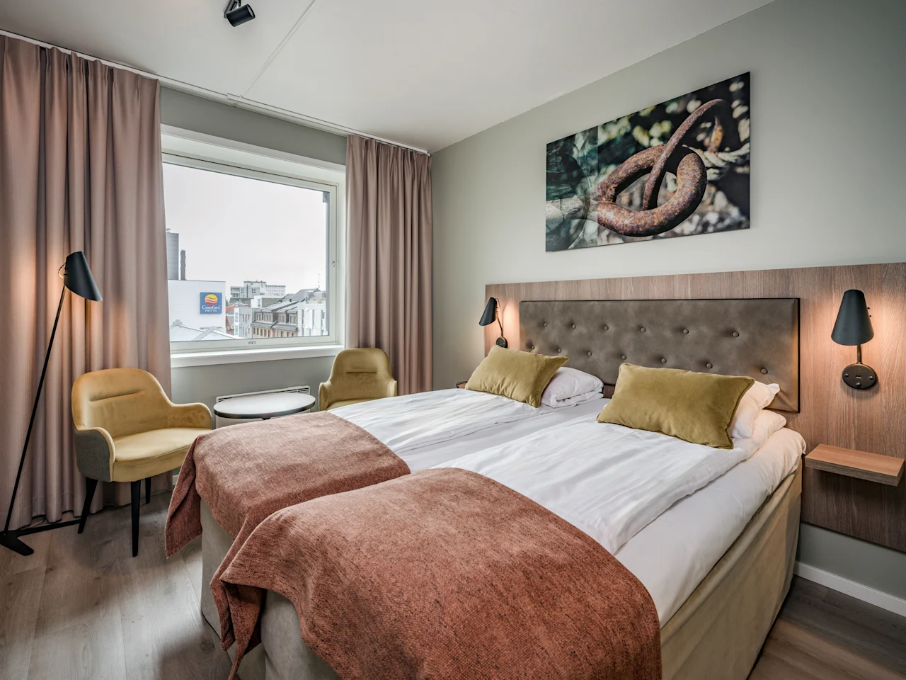 Hotellihuone, jossa on kaksi sänkyä, kaksi keltaista tuolia ja ikkuna, josta on näkymä kaupunkiin. Suuri abstrakti taideteos roikkuu sänkyjen yläpuolella.