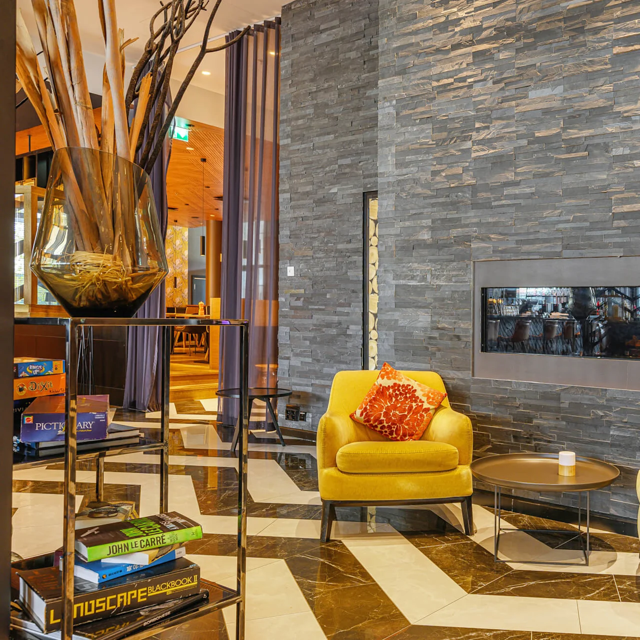 Yellow armchairs with orange pillows sit by a modern, wall-mounted fireplace on a stone wall. Nearby, a small round table holds a candle, and a shelf displays books and games.