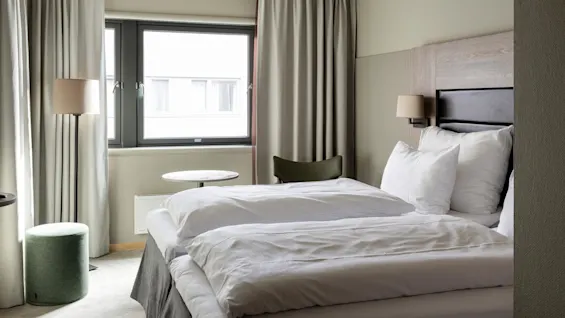A neatly made bed with white linens is in a modern hotel room, featuring light gray curtains, a window, a green chair, a small round table, and a floor lamp.