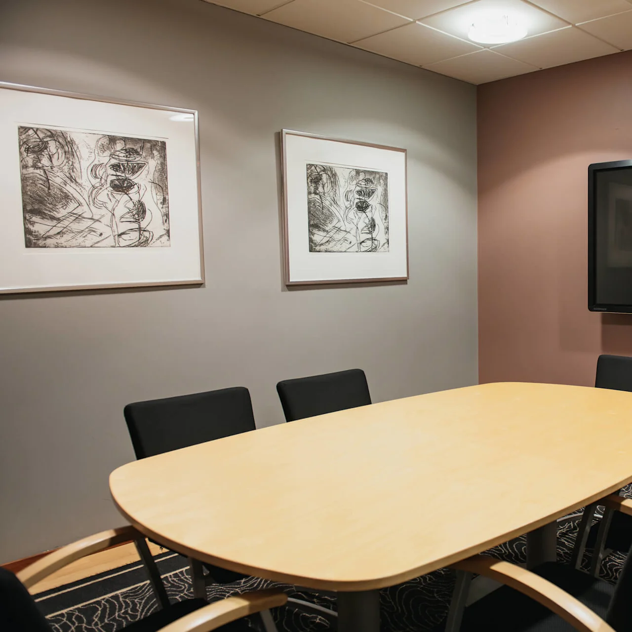 Conference table surrounded by six black chairs; large screen mounted on brown wall; two abstract artworks hang on gray wall. Room is well-lit and carpeted.