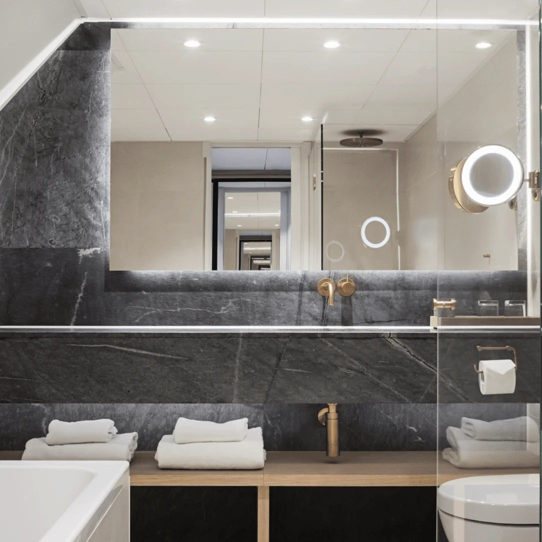 A modern bathroom features a large mirror, illuminated by spotlights, reflecting a continuous hallway. Towels are neatly placed under a dark stone countertop beside a bathtub.