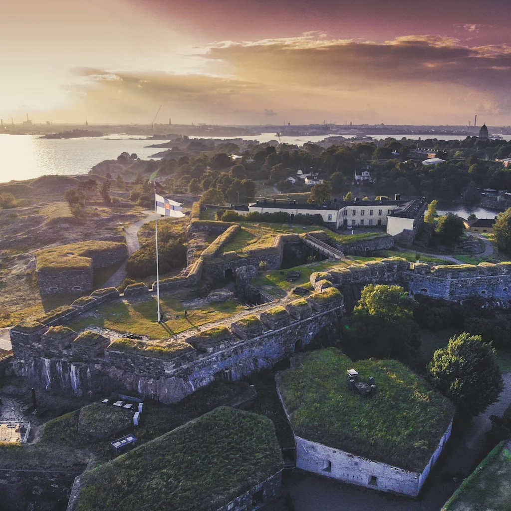 Luftfoto av Sveaborg sjøfestning ved solnedgang, med historiske steinstrukturer og et flagg som vaier over vannet.