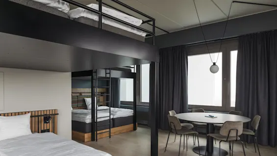 A modern room features a bunk bed and single bed with white linens. A round dining table with chairs is near large windows with dark curtains.