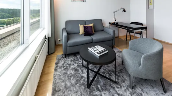 En grå sofa med puter vender mot et rundt svart kaffebord med en bok i et lyst rom med store vinduer og utsikt over grønne trær. Et skrivebord og lampe er i nærheten.
