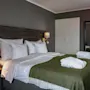 A neatly made bed with white pillows and a green blanket rests against a dark wooden headboard. Wall lamps illuminate the space, with a mirror and nightstand nearby.