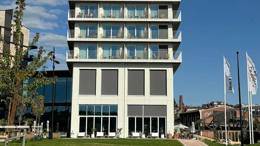 En modern, flervåningsbyggnad står imponerande med balkonger och stora fönster, omgiven av en grön gräsmatta och närliggande träd. Två flaggor lyder "Hotel Riviera" och "Briggen".