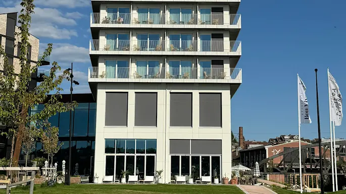 A modern, multi-story building stands tall with balconies and large windows, surrounded by a green lawn and nearby trees. Two flags read "Hotel Riviera" and "Briggen".