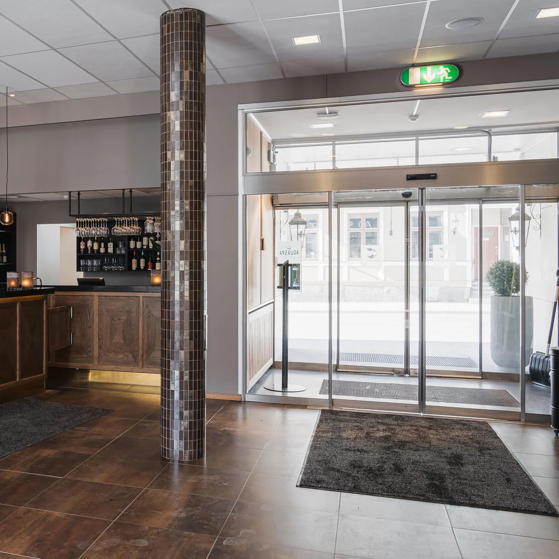 Receptionsdisken är gjord av trä, med hängande lampor ovanför och en vas med en växt. En glasentrédörr visar gatan utanför. Nödutgångsskylt och snacks syns.