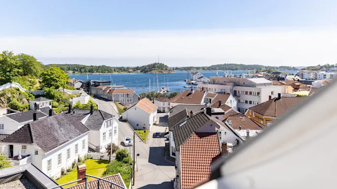 En kystby med hvite hus, røde tak, en gate og en bil, innrammet av et åpent vindu som ser utover en bukt under en klar himmel.