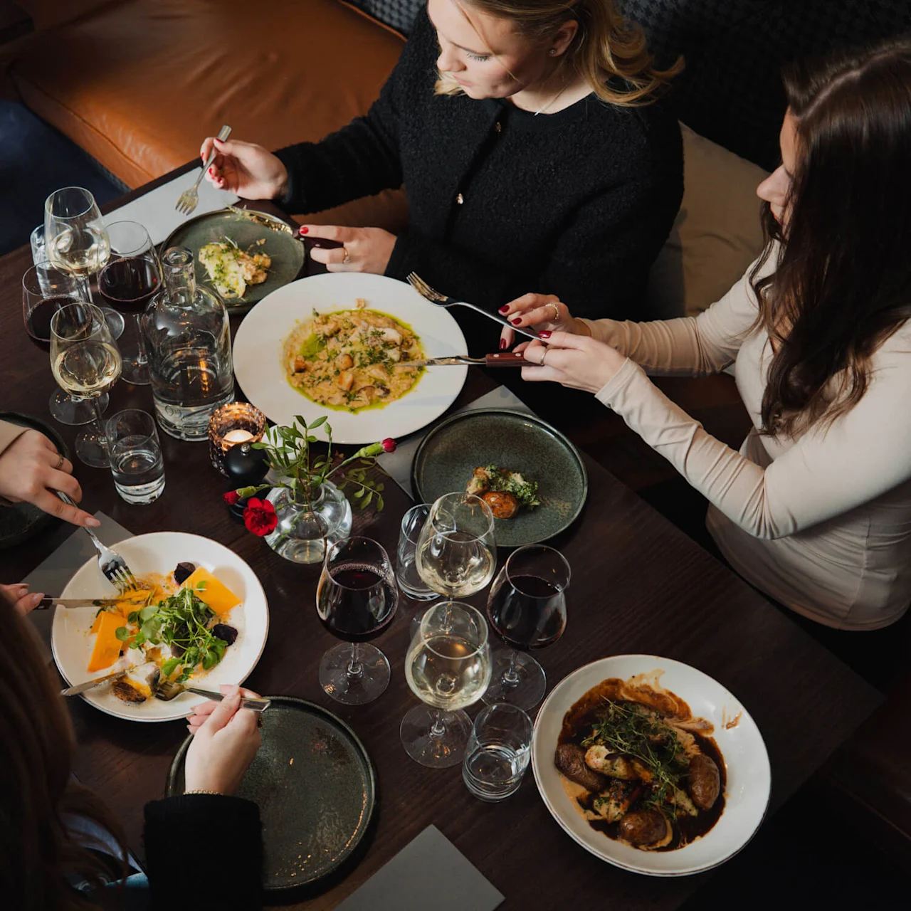 Fire personer spiser forskellige retter med gafler i en restaurationsmiljø. Bordet har flere glas vin og vand blandt dæmpet, hyggelig belysning.