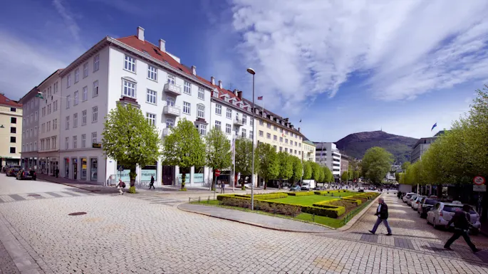 English: A tree-lined street borders a city park with geometric grass patterns; pedestrians walk on the cobblestone path, surrounding tall buildings bearing signage like "THEATRE."