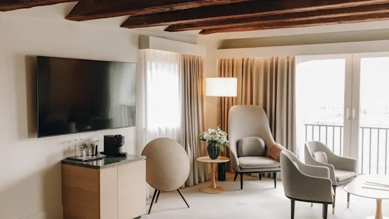 Mysig hotellsvit med synliga takbjälkar i trä, bekväma fåtöljer, en stor TV och balkongdörrar.