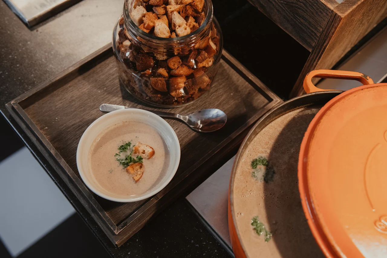 A warm bowl of soup with croutons and fresh herbs, ready to be served from a large pot, with extra croutons available.