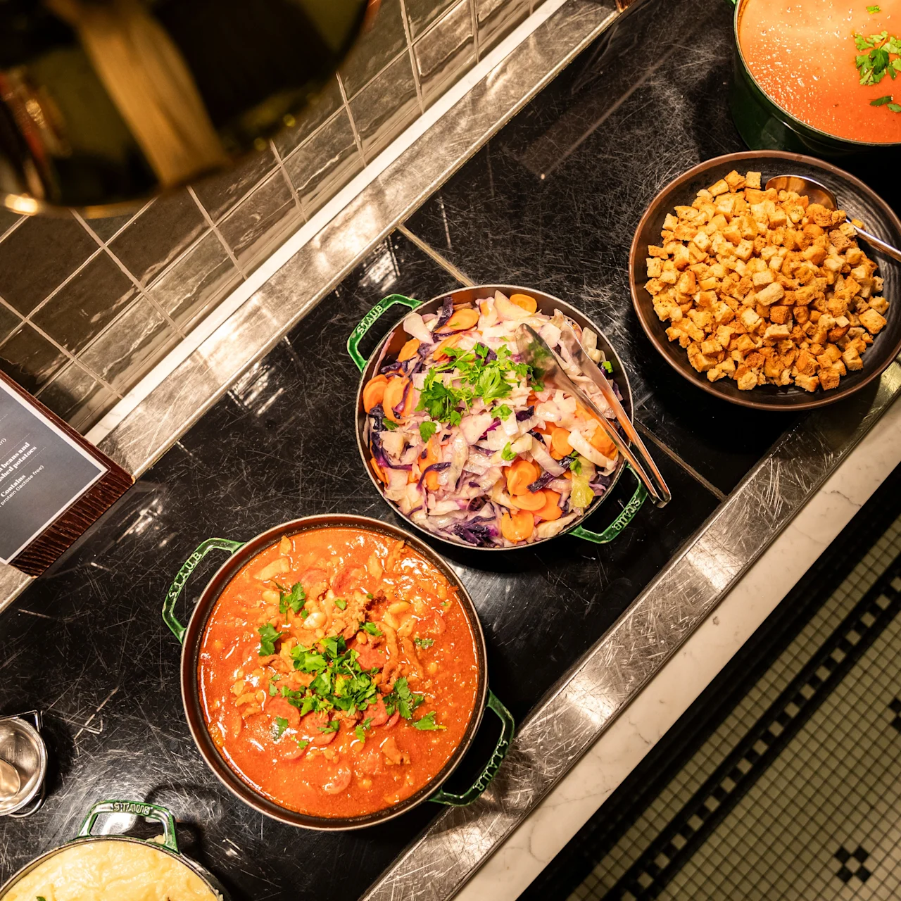 Buffet dishes on a counter; stew garnished with herbs, mixed vegetable salad, cubed tofu. Tiled floor below. Sign: "Vegetarian Buffet - Tomato Soup, Vegetable Stew, Tofu Stir Fry."