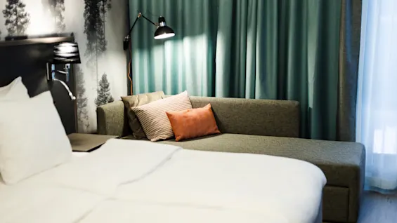 A neatly made bed with white sheets is adjacent to a green sofa, adorned with colorful pillows, set against teal curtains and a wallpaper depicting tall trees.