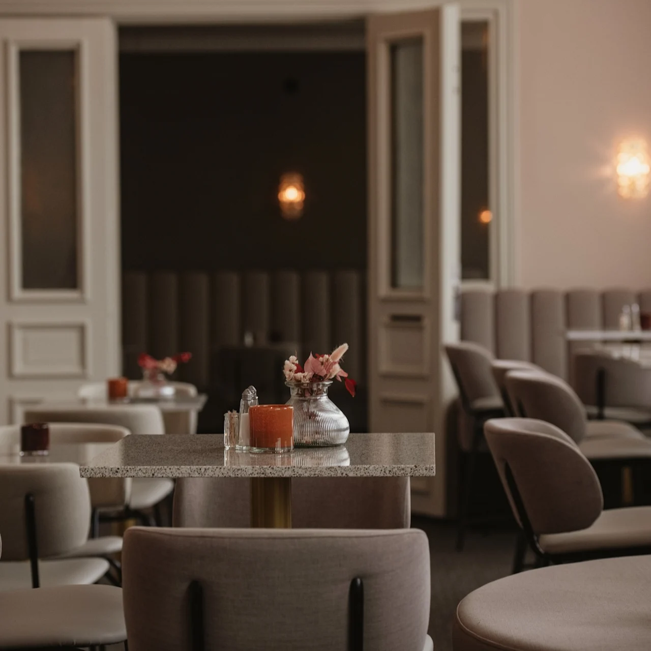 A cozy cafe with beige chairs and tables, some decorated with small flower arrangements, surrounded by soft lighting and partially open double doors.