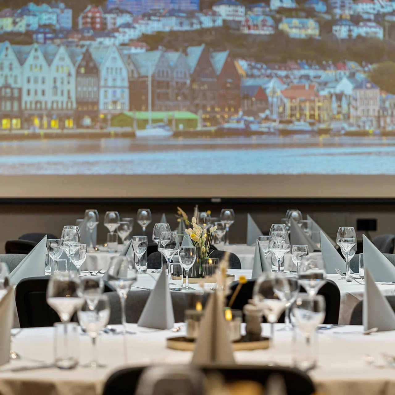 Bordene er elegant dækket med vinglas og servietter i en spisesal, med et stort billede af farverige bygninger ved vandkanten projiceret i baggrunden.