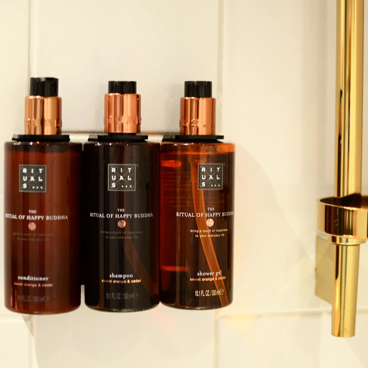 Three brown bottles labeled "The Ritual of Happy Buddha" with a conditioner, shampoo, and shower gel, on a white tiled shelf.
