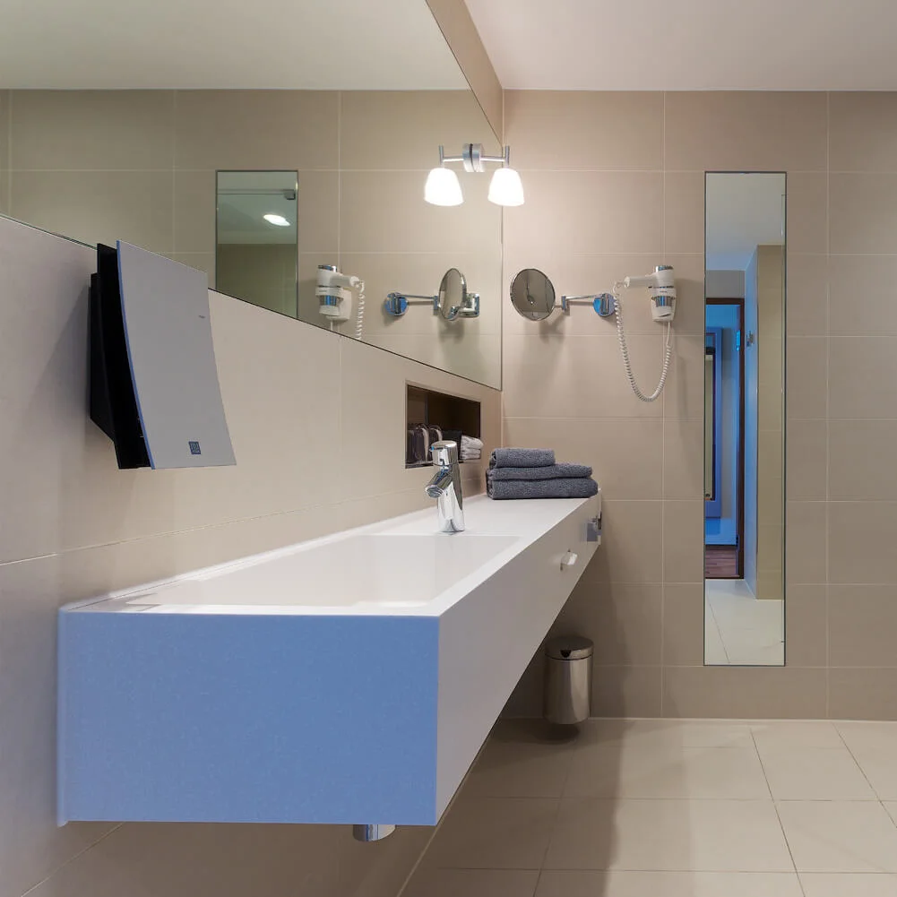 A modern bathroom features a long white sink with a faucet and stacked dark towels, under bright lights. A glass-enclosed shower is on the side with beige tiled walls.