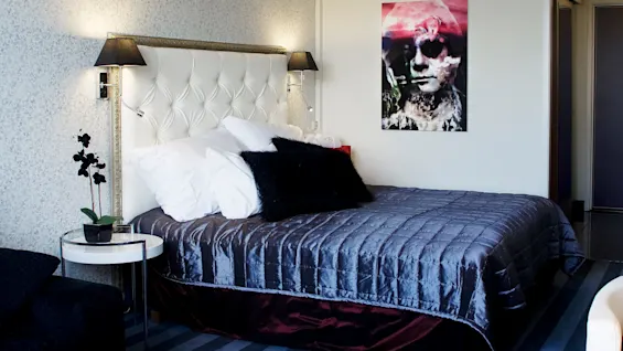A neatly made bed with a white headboard, flanked by lamps, in a room with striped carpet and an abstract picture on the wall.