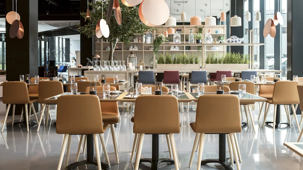 Pöytiä ruskeilla tuoleilla katettu ruokailuun, lasit ja lautasliinat asetettu. Roikkuvat koristevalot, sisäkasvit ja suuret ikkunat lisäävät modernia ravintola-ilmapiiriä.