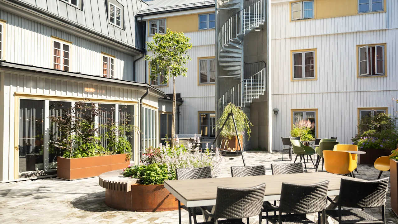 Udendørs gårdhave med spisebord og stole på stenlagt terrasse. Vindeltrappe fører op ad bygningens facade. Plantekasser og små træer pryder området.
