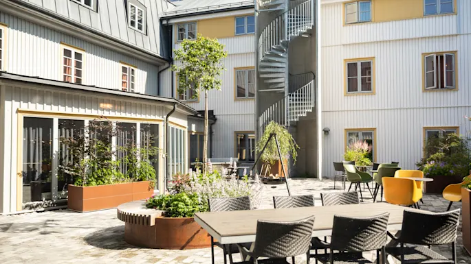 Outdoor courtyard with a dining table and chairs on a stone patio. Spiral staircase leads up the building facade. Potted plants and small trees add greenery to the space.