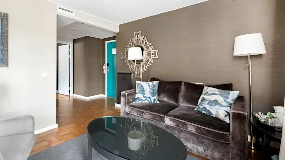 A living room with modern furniture and neutral tones. Dark brown sofa, decorative mirror, and a glass coffee table in a brightly lit space.
