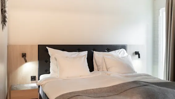 Et komfortabelt hotellrom med en pent oppredd seng, nattbordslamper og et vindu med gardiner, klart for et avslappende opphold.