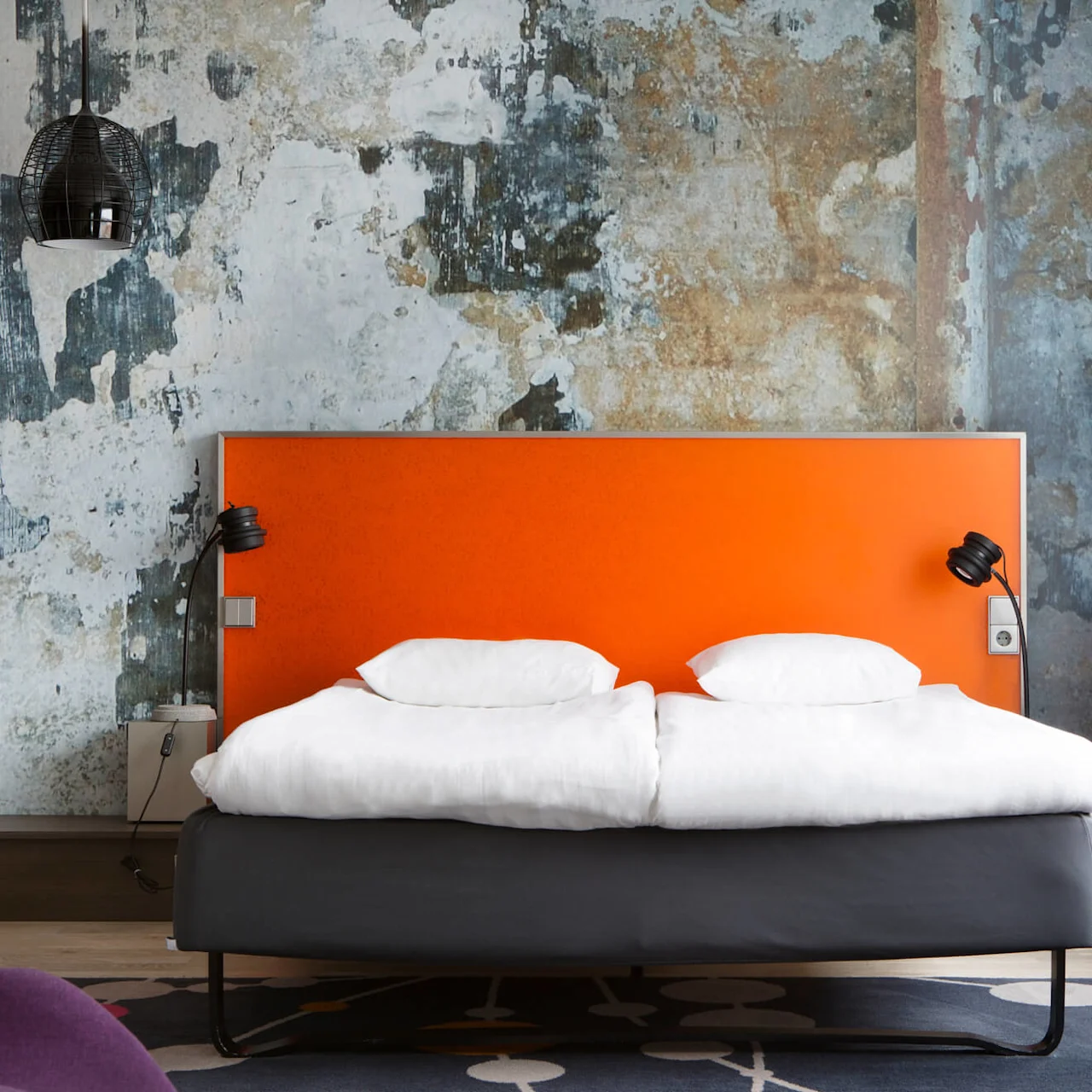 En seng med orange sengegavl står mod en struktureret, slidt væg. To hvide puder ligger på den, omgivet af moderne lamper og et stort vindue med gardin.