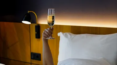 A person lying in bed raises a glass of champagne, with a framed piano keyboard picture overhead.