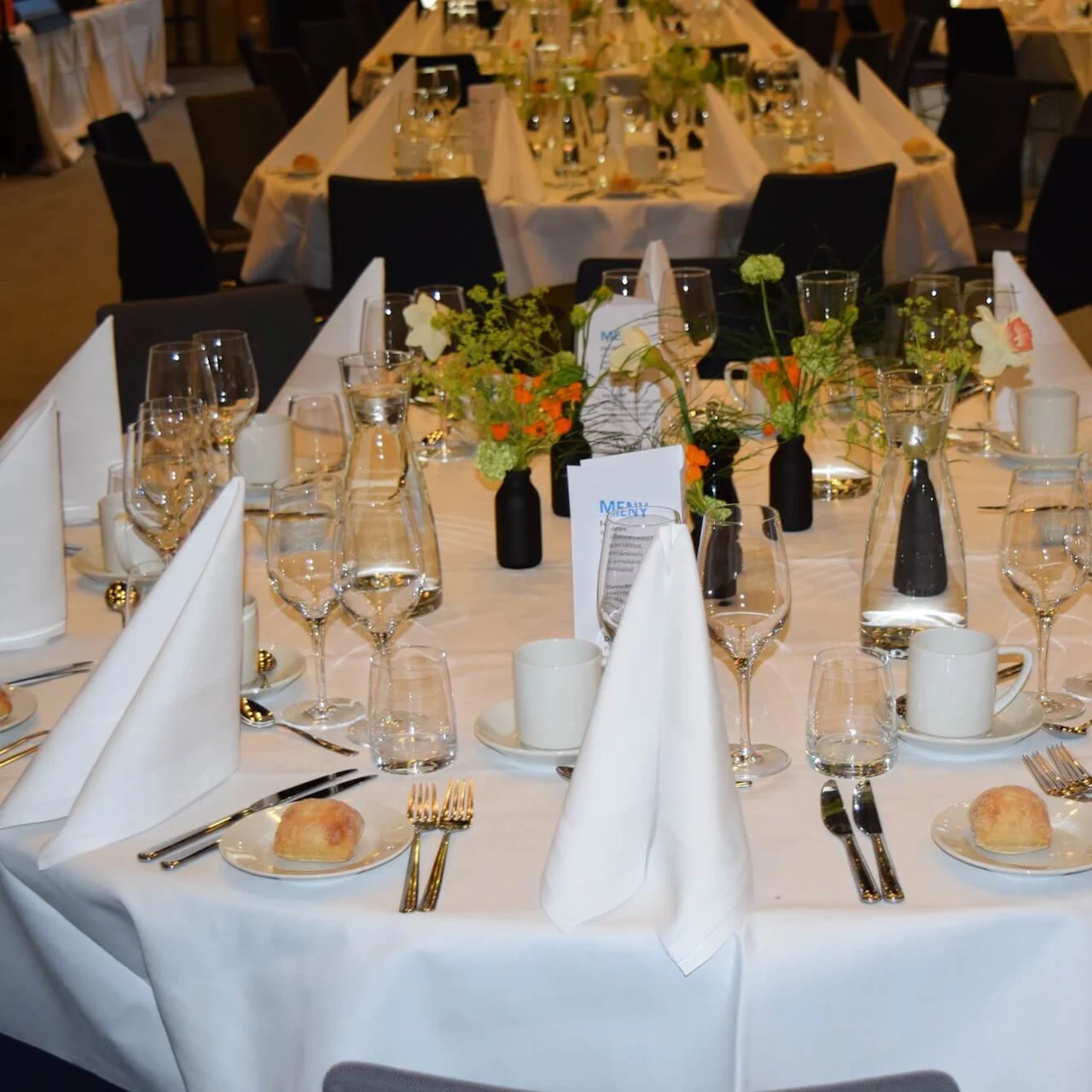Et rundt spisebord er elegant dækket med hvide servietter, glas, menu kort og små blomster i vaser, placeret i en formel banketsal.