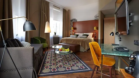 A well-lit hotel room with a bed, chairs, a desk, lamps, and a TV, reflecting a cozy and modern atmosphere.