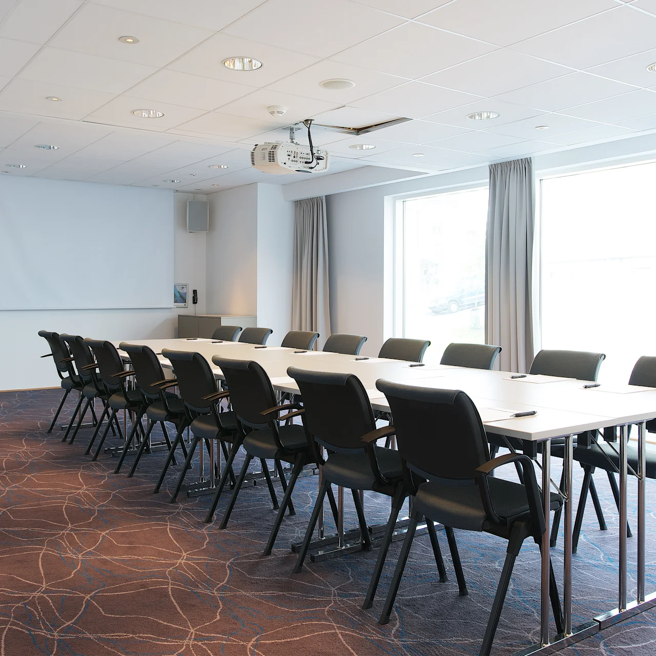 Långt konferensbord omgivet av svarta stolar, placerat i ett rymligt, ljust rum med stora fönster, gardiner och en takmonterad projektor ovanför. En vit projektionsskärm hänger på väggen.