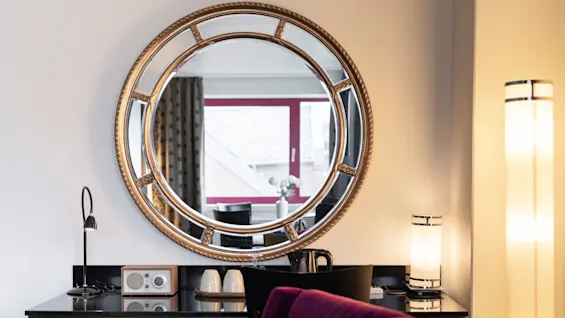 Tyylikäs huone, jossa on suuri pyöreä peili työpöydän yläpuolella lampun, radion ja kuppien kanssa, heijastaen ikkunanäkymää.