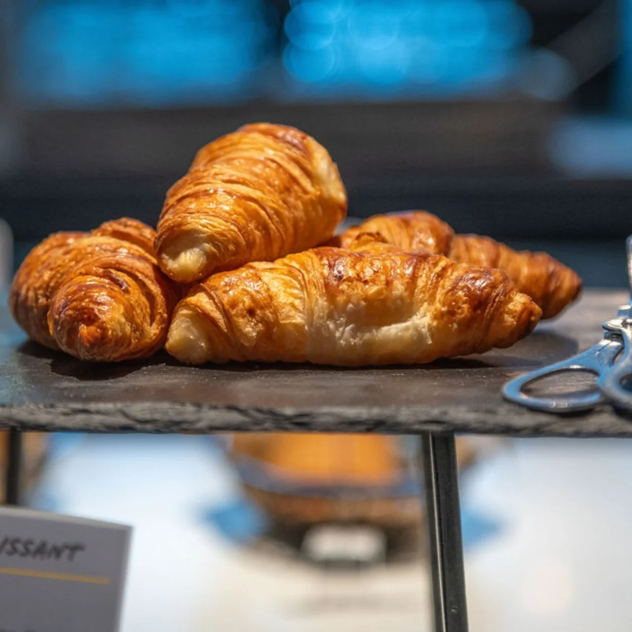 Golden-brown croissants rest on a slate tray with silver tongs beside them; blurred bakery background. 