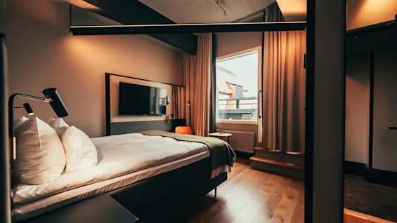 A cozy hotel room with a comfortable bed, a wall-mounted TV, and a window with warm lighting.