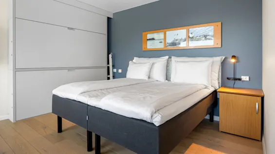 A double bed against a blue wall with a framed picture, side table, and lit lamp in a room.