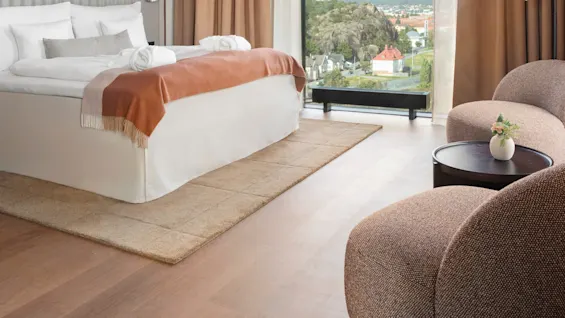 En säng med vita sängkläder och en orange filt vetter mot två stolar och ett litet bord, beläget i ett rum med stora fönster och beige gardiner, som erbjuder en naturskön utsikt.