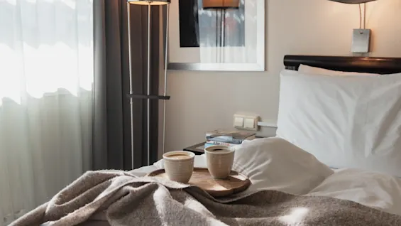 Två koppar kaffe på en bricka i en säng med en mysig filt, vilket antyder en avkopplande morgon eller rumsservice.