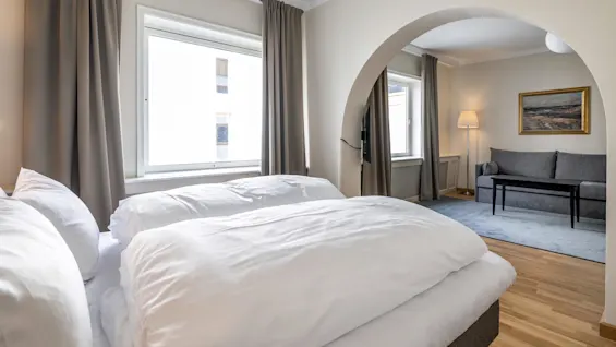 Seng med hvidt sengetøj i forgrunden, under et vindue med beige gardiner. Buegang fører ind til siddeområde med grå sofa, lampe og maleri på væggen.