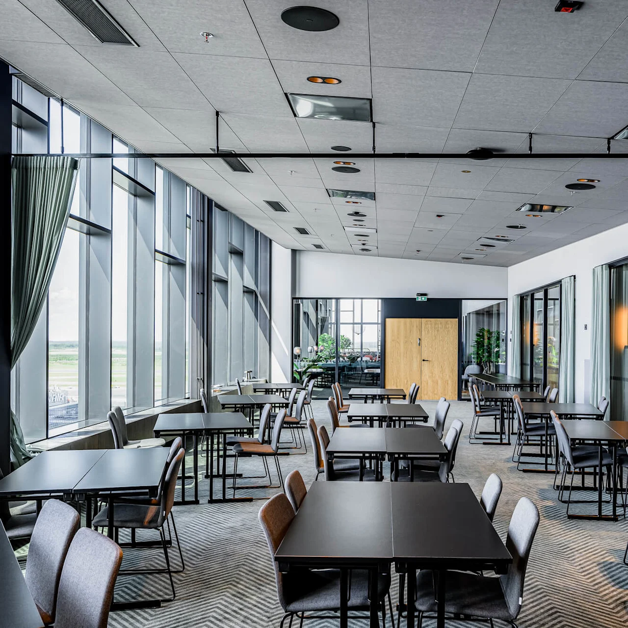 Spacious room with black tables and gray chairs arranged neatly, surrounded by large windows and green curtains, creating a modern and airy environment.