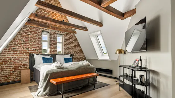 Et hyggeligt lofts-soveværelse har en seng med gråt sengetøj og blå puder, en orange bænk, murstensvæg, træbjælker, skråt loft og et vægmonteret TV ved siden af et konsolbord.