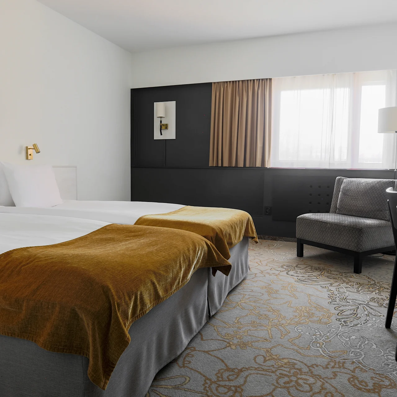 A neatly made bed with brown throws, sits in a minimalistic hotel room with white walls, a gray armchair, and a black desk next to a large window.