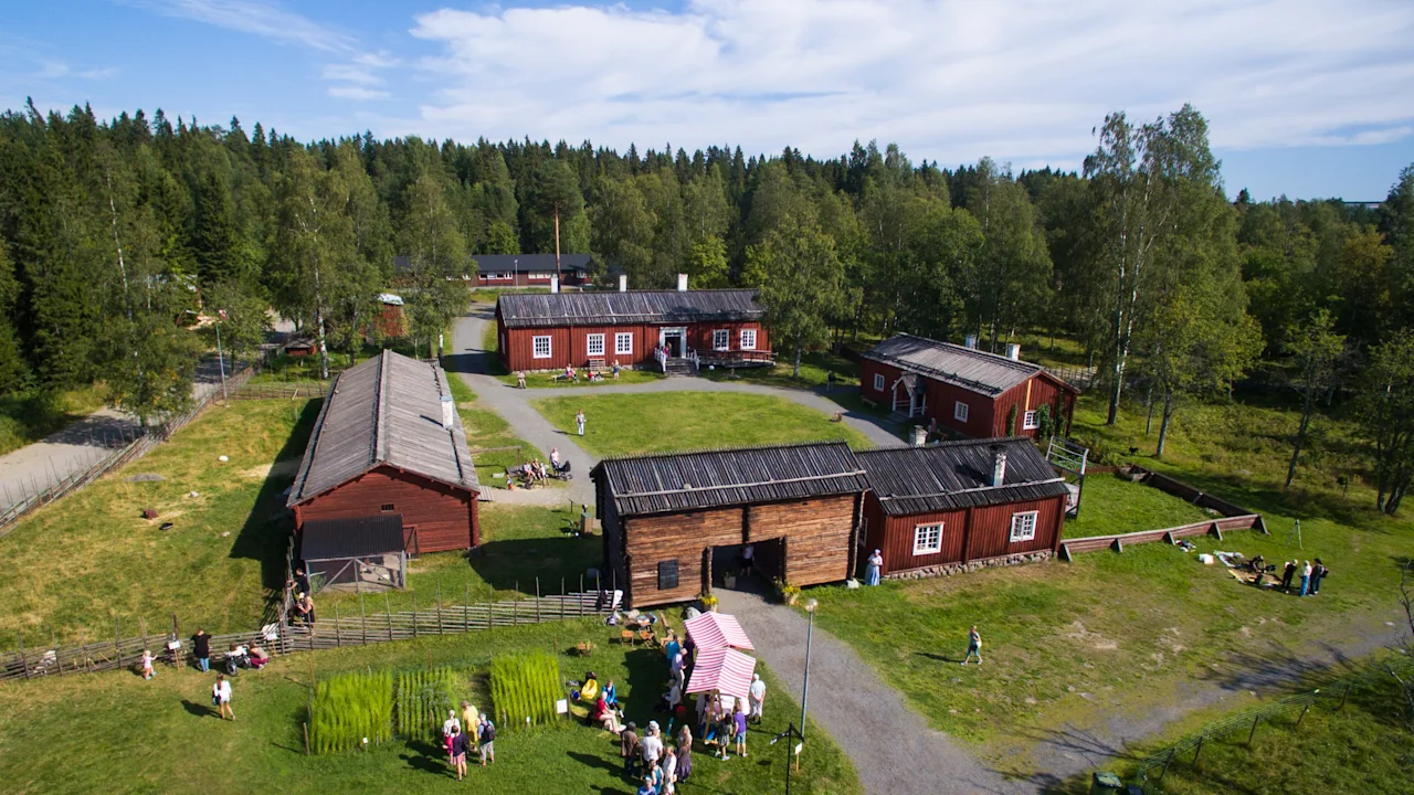 An aerial view of a historical village with red and wooden buildings, surrounded by green grass and trees. People are enjoying an outdoor event.