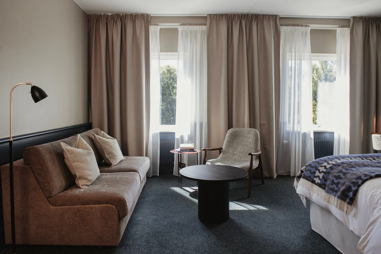 Ett bekvämt rum med en soffa, en fåtölj och en säng, upplyst av solljus som strömmar in genom draperade fönster.