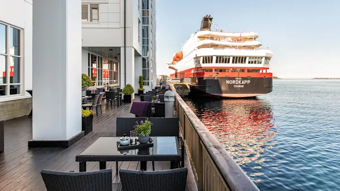 A large ship labeled "NORDKAPP TROMSØ" is docked beside a modern waterfront building. Outdoor seating with tables and plants lines a wooden deck.