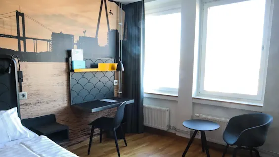 Ett hotellrum med en enkelsäng, skrivbord, stol och två fönster. En väggmålning visar en stadssilhuett och bro. Ljust trägolv och enkel inredning.